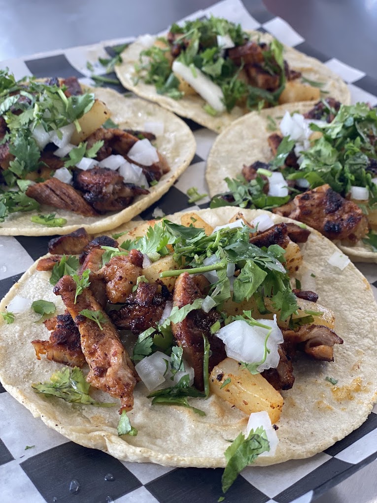 Four al pastor tacos with pineapple, onion and cilantro on checkered paper